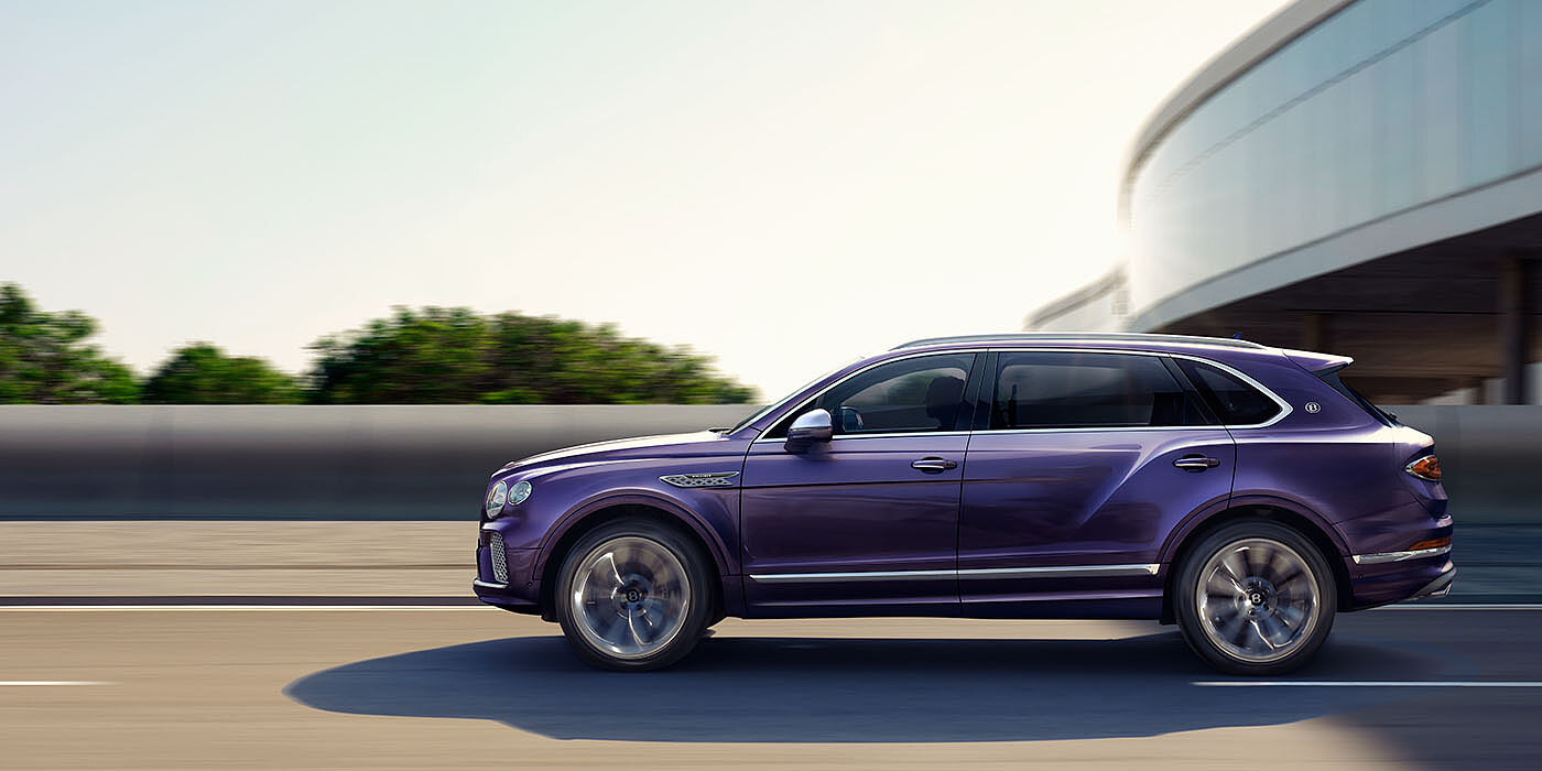 Bentley High Wycombe Bentley Bentayga Extended Wheelbase Mulliner luxury SUV profile in Tanzanite Purple paint, driving on an country road past trees.